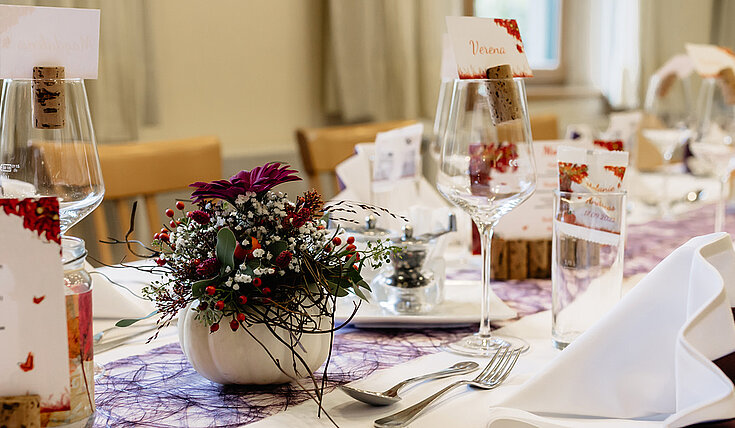 Auf einer Tafel wurde weiß eingedeckt. In den weißen Mundservietten wurden violette Servietten eingearbeitet. In der Tischmitte verläuft ein violetter Tischläufer. In kleinen weißen Kürbissen wurden Blumen eingearbeitet.
