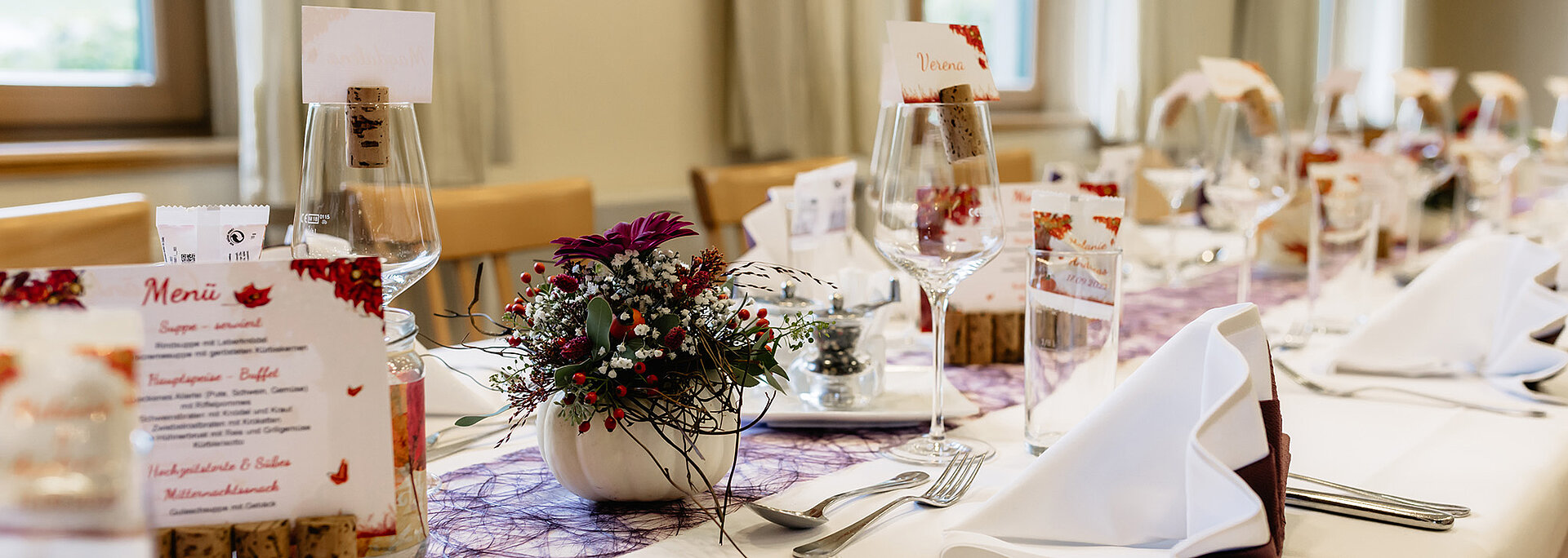 Auf einer Tafel wurde weiß eingedeckt. In den weißen Mundservietten wurden violette Servietten eingearbeitet. In der Tischmitte verläuft ein violetter Tischläufer. In kleinen weißen Kürbissen wurden Blumen eingearbeitet.