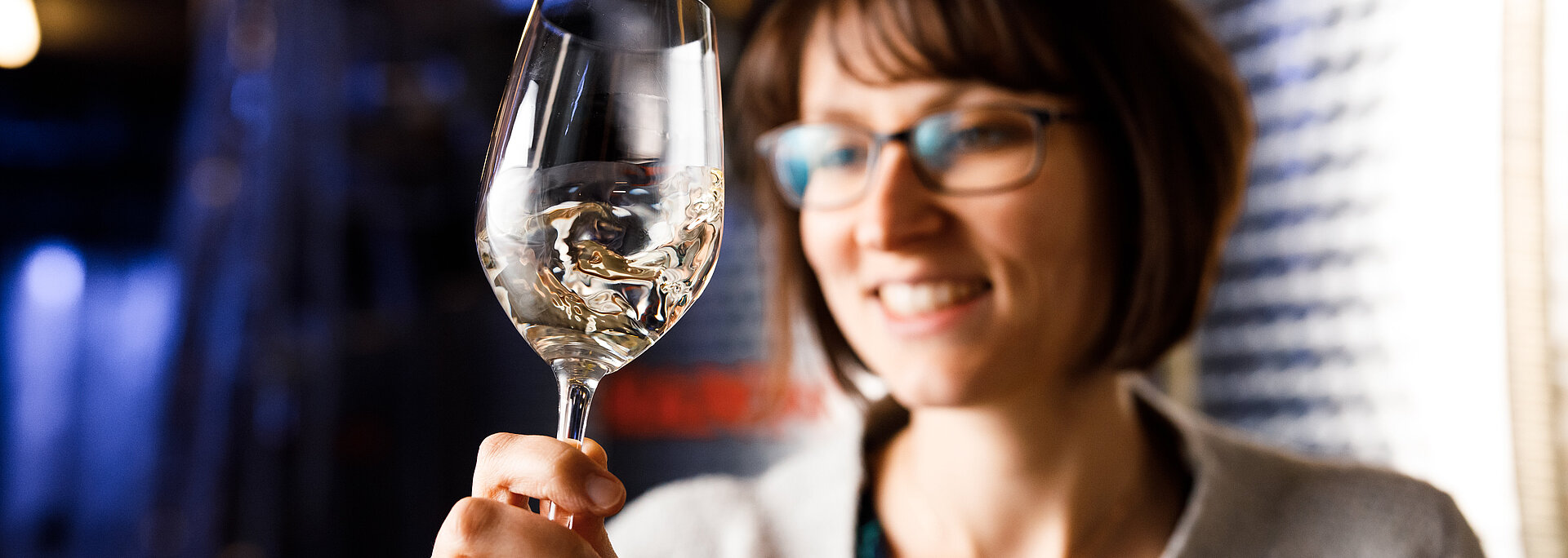 Monika Neustifter steht im Weinkeller vor einem Stahltank und schwenkt ein Wein im Weißweinglas. Der Fokus liegt auf dem Weinglas, während Monika verschwommen im Hintergrund steht.