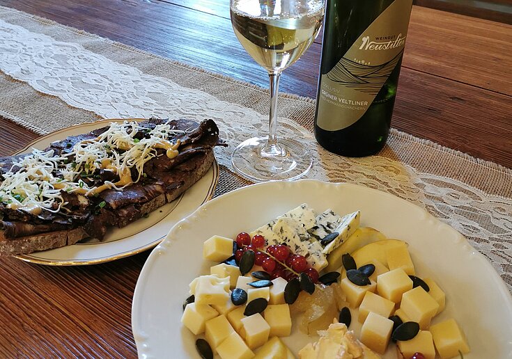 Auf dem Tisch ist ein weißes Teller mit verschiedenen Käsesorten, die klein geschnitten sind. Neben dem Käseteller ist ein Weiteres mit einem Brot mit Blunzen, Kren und Senf. Dahinter steht ein Glas mit Weißwein, sowie eine Weißweinflasche.