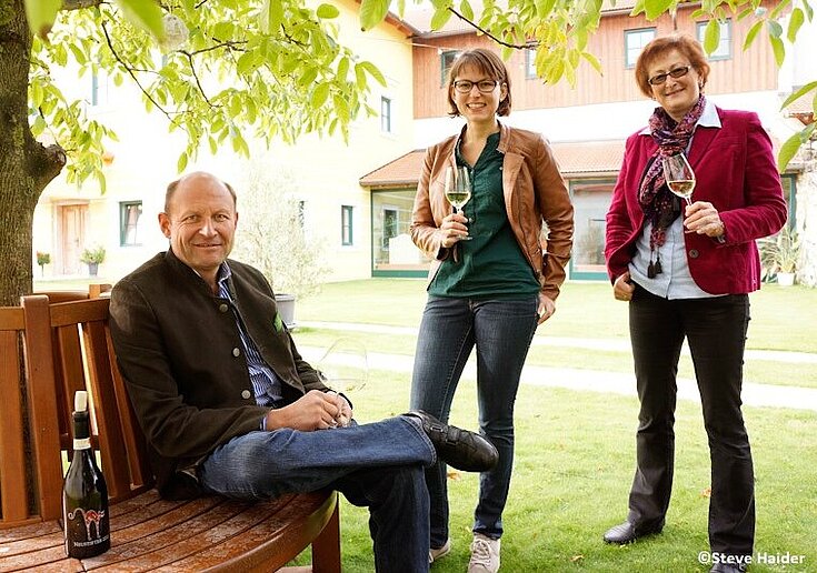 Karl Neustifter sitzt mit einem Glas Wein unter einem Baum auf einer Bank. Auf der Bank steht auch eine Weinflasche. Neben ihm stehen Monika Neustifter und Brigitte Neustifter ebenfalls in der Hand ein Glas Wein.